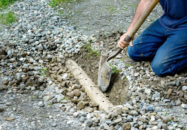 Original photograph: a perforated French drain pipe being set into a trench surrounded by drainage gravel, a worker shoveling stone in to encase the pipe